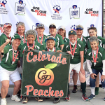A women's softball team, aged 79-91, smiling together for a team picture outside in their green and white uniforms and red medals, with the center of the front row holding a Colorado Peaches banner, all in front of a white picture banner with assorted logos likely of tournament sponsors.